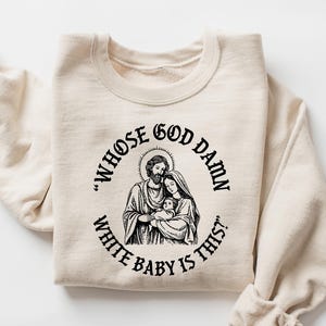May include: A cream-coloured sweatshirt featuring a black graphic of a religious family and the text "WHOSE GOD DAMN WHITE BABY IS THIS?" The sweatshirt is made of a soft material and has long sleeves.