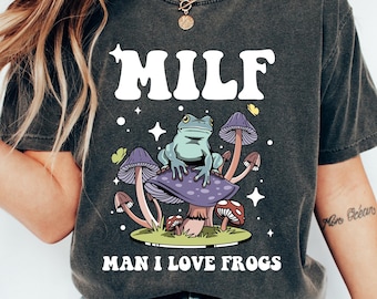 May include: A dark gray t-shirt with the text "MILF MAN I LOVE FROGS" in white. A cartoon frog sits on a purple mushroom with other mushrooms in the background.