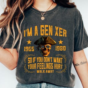 May include: Dark grey t-shirt with the text "I'M A GEN XER" and the years 1965 and 1980. The shirt features a skull wearing a pirate hat. The text "SO IF YOU DON'T WANT YOUR FEELINGS HURT WALK AWAY" is also printed on the shirt.