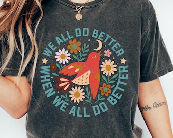 May include: Dark gray t-shirt with a circular design featuring a red bird, flowers, and the text "WE ALL DO BETTER WHEN WE ALL DO BETTER." The shirt is paired with blue jeans and white sneakers. The overall theme is a positive message.