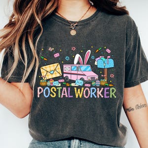 May include: Dark gray t-shirt with a colorful "Postal Worker" graphic. The design features a pink mail truck with bunny ears, envelopes, and Easter eggs. The shirt is a comfortable, casual style.