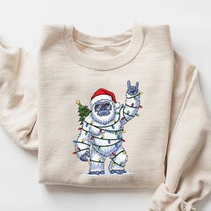 May include: A cream-colored sweatshirt featuring a festive yeti design. The yeti is adorned with Christmas lights, a Santa hat, and a small Christmas tree. The yeti is making a rock-and-roll hand gesture. The sweatshirt is folded.