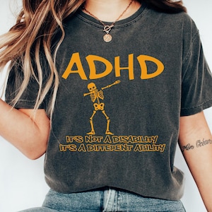 May include: A dark grey t-shirt with the text "ADHD" in yellow. A yellow skeleton is doing the dab dance. The text below the skeleton reads "It's not a disability it's a different ability".