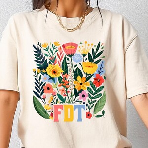 Puede incluir: Una camiseta de color crema con un diseño floral y el texto "FDT" en letras de colores.