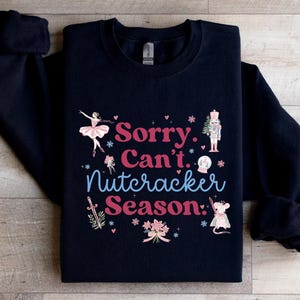 May include: A navy blue sweatshirt with the text "Sorry, Can't, Nutcracker Season" in pink and blue. The design includes illustrations of a ballerina, nutcracker, and mouse, with floral and star accents.