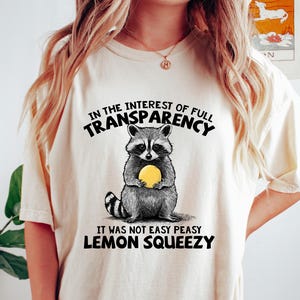 May include: Cream-colored t-shirt featuring a raccoon holding a yellow lemon. The text on the shirt reads "IN THE INTEREST OF FULL TRANSPARENCY, IT WAS NOT EASY PEASY LEMON SQUEEZY". The shirt is a casual wear item.