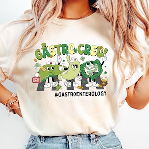 May include: Cream-colored t-shirt with a graphic featuring cartoon organs on a road, the text "Gastro Crew!" and the hashtag #Gastroenterology. The design includes green and white colors. The shirt is paired with light blue jeans.