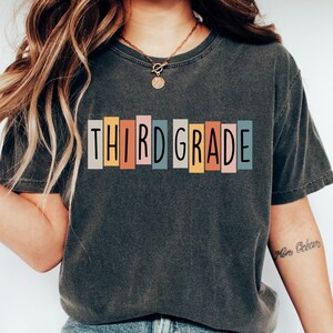 Op de afbeelding: Donkergrijs T-shirt met de woorden "THIRD GRADE" in kleurrijke blokletters. Het shirt wordt gedragen met een blauwe spijkerbroek en witte sneakers. Het algemene thema is schoolkleding.