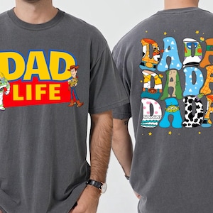 K&ouml;nnte beinhalten: Dunkelgraues T-Shirt mit "DAD LIFE" in Gelb und Rot, mit Buzz Lightyear und Woody. Auf der R&uuml;ckseite des Shirts steht "DADA DADA" in bunten, cartoonartigen Buchstaben. Ein lustiges, thematisches Kleidungsst&uuml;ck.