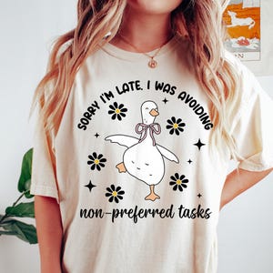 Peut inclure: T-shirt crème avec un dessin fantaisiste d'une oie de dessin animé avec un nœud papillon rose, entourée de marguerites noires et d'étoiles. Le texte dit : « SORRY I'M LATE, I WAS AVOIDING non-preferred tasks. »