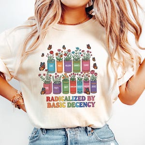Pode incluir: Camiseta creme com um gráfico colorido de vasos de flores, borboletas e o texto "RADICALIZED BY BASIC DECENCY". Os vasos de flores são rotulados com palavras como "AMOR", "PAZ" e "UNIDADE". A camiseta é combinada com jeans azuis.