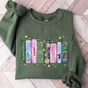 Pu&ograve; includere: Una felpa verde oliva con una grafica colorata di dorsi di libri. I titoli dei libri includono "The Wizard and I", "Defying Gravity", "Popular", "I've Had So Many Friends But Only One That Mattered" e "Pink Goes Green".