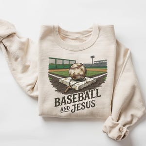 Könnte beinhalten: Ein cremefarbenes Sweatshirt mit Rundhalsausschnitt und einer grafischen Illustration eines Baseballs, der auf einer Home Plate in einem Stadion liegt. Ein kleines Holzkreuz hängt am Baseball. Der Text "BASEBALL AND JESUS" steht darunter.
