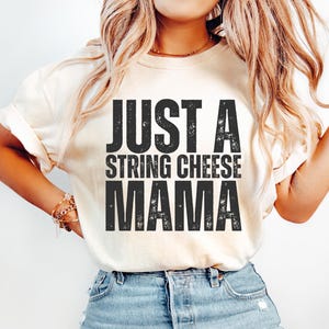 Peut inclure: T-shirt crème avec l'inscription "JUST A STRING CHEESE MAMA" en lettres noires délavées. Le t-shirt est associé à un jean en denim bleu clair. Le style est décontracté et confortable.