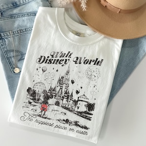 Puede incluir: Camiseta blanca con un gr&aacute;fico negro del castillo de Walt Disney World, Mickey Mouse y el texto "The happiest place on earth". La camiseta se combina con vaqueros azul claro y un sombrero beige.