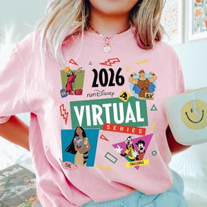May include: A light pink t-shirt with the text "2026 runDisney VIRTUAL SERIES" and cartoon character graphics. The shirt features images of Hercules, Pocahontas, and Goofy. A white mug with a yellow smiley face is held in the hand.