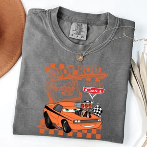 May include: Grey t-shirt with an orange cartoon car design, featuring the text "Shoot Rod" and "Cars". The design includes checkered flags and a vintage car illustration. The shirt is a Comfort Colors brand.