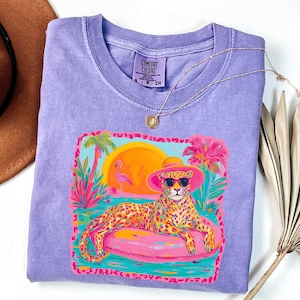 May include: A purple Comfort Colors t-shirt featuring a colorful graphic of a leopard wearing sunglasses and a hat, lounging on a pink inflatable ring. The graphic includes a sunset, palm trees, and a flamingo. A gold necklace is draped across the shirt.