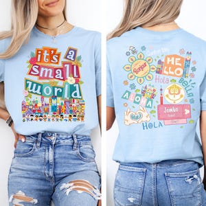 K&ouml;nnte beinhalten: Hellblaues T-Shirt mit dem Text "it's a small world" in bunten Buchstaben und einer Grafik einer skurrilen Stadtlandschaft. Auf der R&uuml;ckseite des Shirts befinden sich verschiedene Gr&uuml;&szlig;e in verschiedenen Sprachen, darunter "Bonjour", "Hola" und "Aloha".