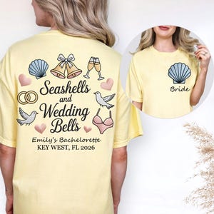 以下が含まれることがあります： 貝殻と結婚をテーマにした、薄い黄色のTシャツ。Tシャツの背面には、貝殻、結婚式の鐘、シャンパングラス、ハート、鳩、ブラジャーのイラストが描かれています。「Seashells and Wedding Bells」と「Emily's Bachelorette KEY WEST, FL 2026」の文字。