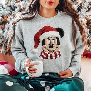 May include: A light gray crewneck sweatshirt featuring a large embroidered graphic of Minnie Mouse wearing a red and white Santa hat and a matching red and white striped scarf. The design is centered on the chest of the long-sleeved garment.