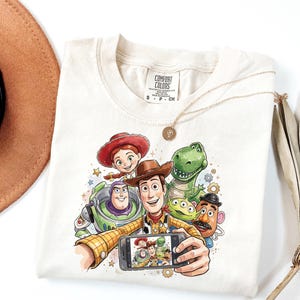 Może przedstawiać: Kremowa koszulka z kolorową grafiką postaci z Toy Story robiących sobie selfie. Projekt zawiera Woody'ego, Buzz'a Astrala, Jessie, Rexa i inne postacie. Koszulka ma metkę Comfort Colors.