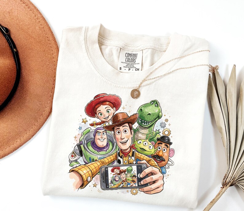 Może przedstawiać: Kremowa koszulka z kolorową grafiką postaci z Toy Story robiących sobie selfie. Projekt zawiera Woody'ego, Buzz'a Astrala, Jessie, Rexa i inne postacie. Koszulka ma metkę Comfort Colors.