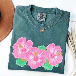 May include: A dark teal short-sleeve t-shirt featuring a graphic of three large pink hibiscus flowers with green leaves. The shirt has a crew neckline and a Comfort Colors brand label on the inside collar indicating a size small.