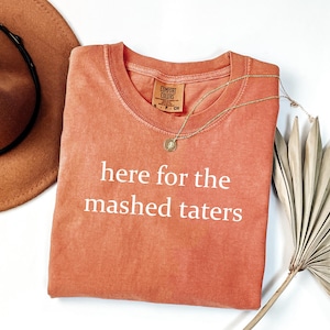 Puede incluir: Una camiseta de color coral con el texto "here for the mashed taters" en blanco. Un collar dorado con un colgante está sobre la camiseta. Un sombrero marrón está en la esquina superior izquierda.