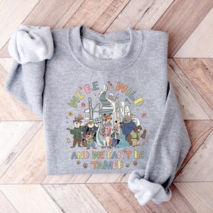 Puede incluir: Una sudadera gris con el texto "WE'RE WILD AND WE CAN'T BE TAMED" en letras coloridas. El gr&aacute;fico presenta personajes de dibujos animados de animales frente a un horizonte urbano. Las mangas est&aacute;n enrolladas.