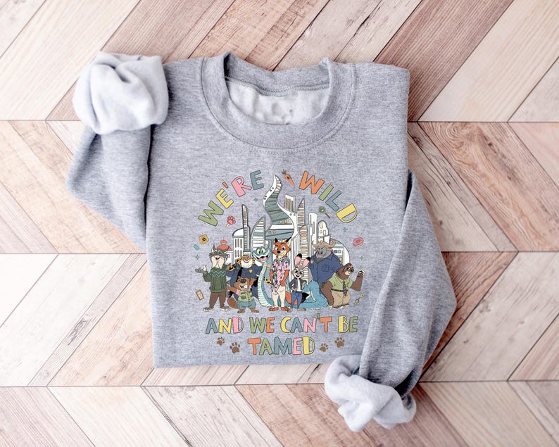 Puede incluir: Una sudadera gris con el texto "WE'RE WILD AND WE CAN'T BE TAMED" en letras coloridas. El gr&aacute;fico presenta personajes de dibujos animados de animales frente a un horizonte urbano. Las mangas est&aacute;n enrolladas.