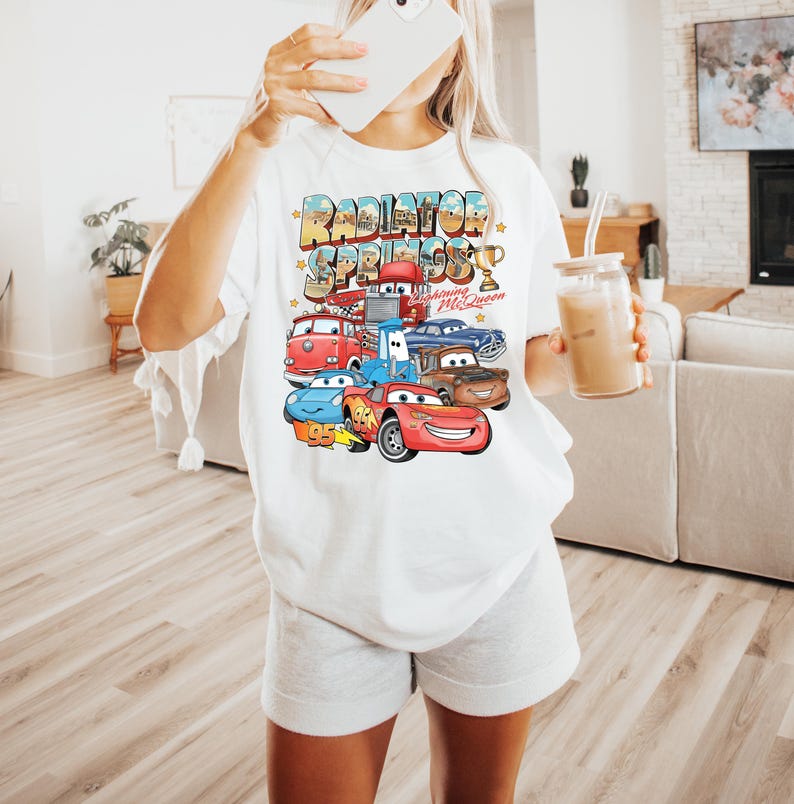 Puede incluir: Camiseta blanca con un gr&aacute;fico colorido de personajes de la pel&iacute;cula Cars, incluyendo a Rayo McQueen. El texto "Radiator Springs" se muestra encima de los personajes. La camiseta es de estilo casual y oversize.