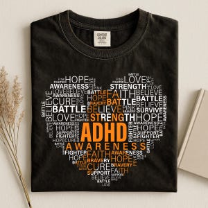 Op de afbeelding: Een zwart t-shirt met ronde hals en een hartvormige afbeelding bestaande uit diverse woorden zoals ADHD, AWARENESS, HOPE, BATTLE, STRENGTH, FAITH, BELIEVE, CURE, BRAVERY, SUPPORT, FIGHTER, SURVIVE, LOVE en PINK in witte en oranje tekst.