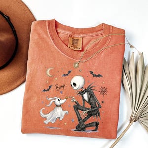 May include: A burnt orange t-shirt featuring a graphic of a skeletal character in a pinstripe suit kneeling to touch the nose of a ghost dog. The design includes the text Boop, a crescent moon, bats, stars, and a spiderweb. The shirt has a crew neck.
