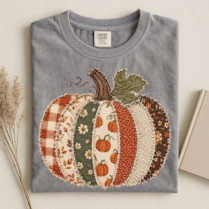 May include: A slate blue crew neck t-shirt featuring a large, central pumpkin graphic composed of various fabric-like patches. The pumpkin design includes patterns of orange gingham, autumn leaves, white daisies on green, and brown floral prints.