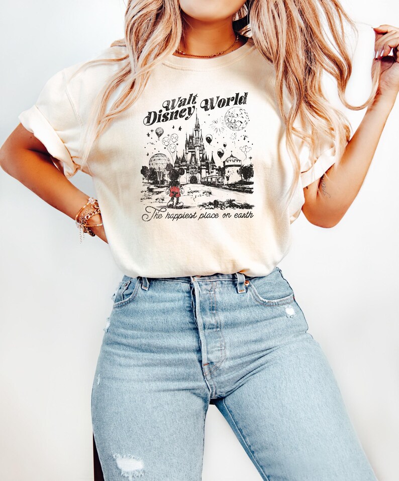 Puede incluir: Camiseta color crema con un gr&aacute;fico en blanco y negro del castillo de Cenicienta, globos y el texto "Walt Disney World" y "The happiest place on earth." La camiseta se combina con vaqueros azul claro.