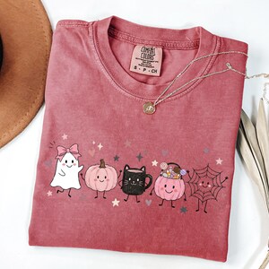 May include: A dusty rose crewneck t-shirt featuring a graphic print of a white ghost with a pink bow, a pink pumpkin, a black cat mug, a pink pumpkin bucket filled with candy, and a spider web, all with small stars and hearts.
