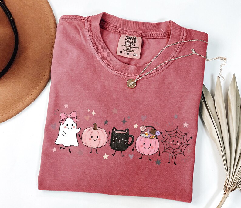 May include: A dusty rose crewneck t-shirt featuring a graphic print of a white ghost with a pink bow, a pink pumpkin, a black cat mug, a pink pumpkin bucket filled with candy, and a spider web, all with small stars and hearts.