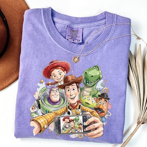 Może przedstawiać: Lawendowa koszulka Comfort Colors z grafiką postaci z filmu Toy Story robiących sobie selfie. Postacie to Woody, Buzz Lightyear, Jessie, Rex i Pan Bulwa. Złoty naszyjnik jest zarzucony na koszulkę.