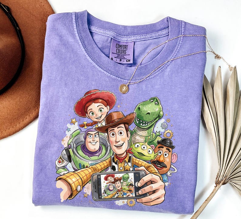 Może przedstawiać: Lawendowa koszulka Comfort Colors z grafiką postaci z filmu Toy Story robiących sobie selfie. Postacie to Woody, Buzz Lightyear, Jessie, Rex i Pan Bulwa. Złoty naszyjnik jest zarzucony na koszulkę.