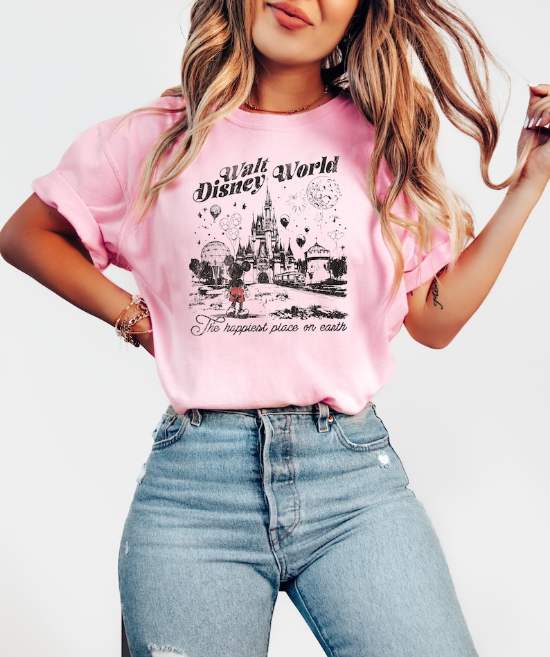 Puede incluir: Camiseta rosa claro con un gr&aacute;fico en blanco y negro del castillo de Walt Disney World, Mickey Mouse y otros elementos del parque. Tambi&eacute;n se muestra el texto "Walt Disney World" y "The happiest place on earth".