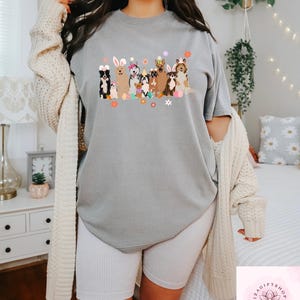 May include: A gray t-shirt with a graphic of dogs wearing bunny ears and holding Easter eggs. The dogs are surrounded by flowers. The t-shirt is being worn by a person.