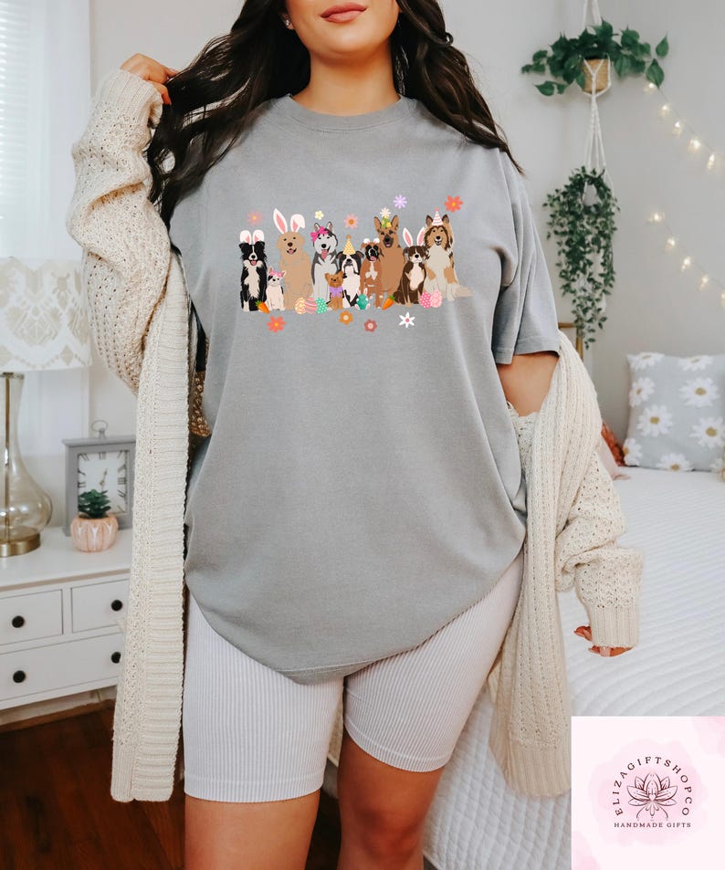 May include: A gray t-shirt with a graphic of dogs wearing bunny ears and holding Easter eggs. The dogs are surrounded by flowers. The t-shirt is being worn by a person.