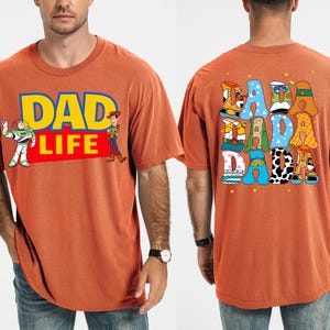 K&ouml;nnte beinhalten: Gebrannt orangefarbenes T-Shirt mit dem Text "DAD LIFE" in Gelb und Blau, mit den Zeichentrickfiguren Buzz Lightyear und Woody. Auf der R&uuml;ckseite des Shirts steht das Wort "DADA" in bunten, cartoonartigen Buchstaben. Ein l&auml;ssiges, bequemes T-Shirt.