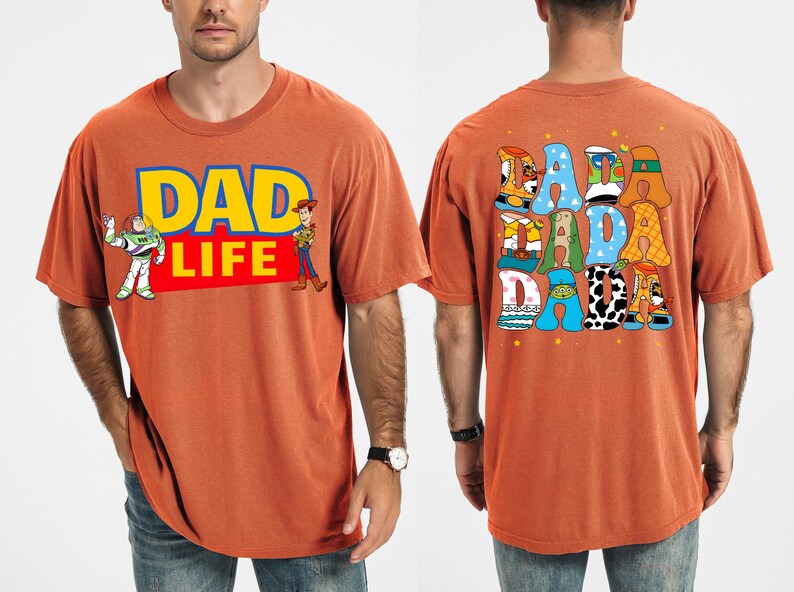 K&ouml;nnte beinhalten: Gebrannt orangefarbenes T-Shirt mit dem Text "DAD LIFE" in Gelb und Blau, mit den Zeichentrickfiguren Buzz Lightyear und Woody. Auf der R&uuml;ckseite des Shirts steht das Wort "DADA" in bunten, cartoonartigen Buchstaben. Ein l&auml;ssiges, bequemes T-Shirt.