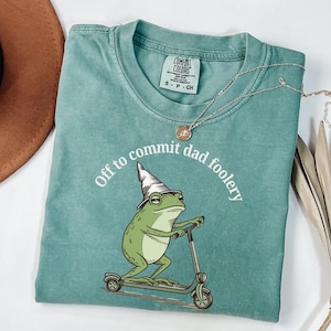 Op de afbeelding: Een turquoise Comfort Colors t-shirt met een cartoon kikker op een scooter, met een tovenaars hoed. De tekst "Off to commit dad foolery" staat boven de kikker. Een zilveren ketting met een kleine hanger ligt over het shirt.