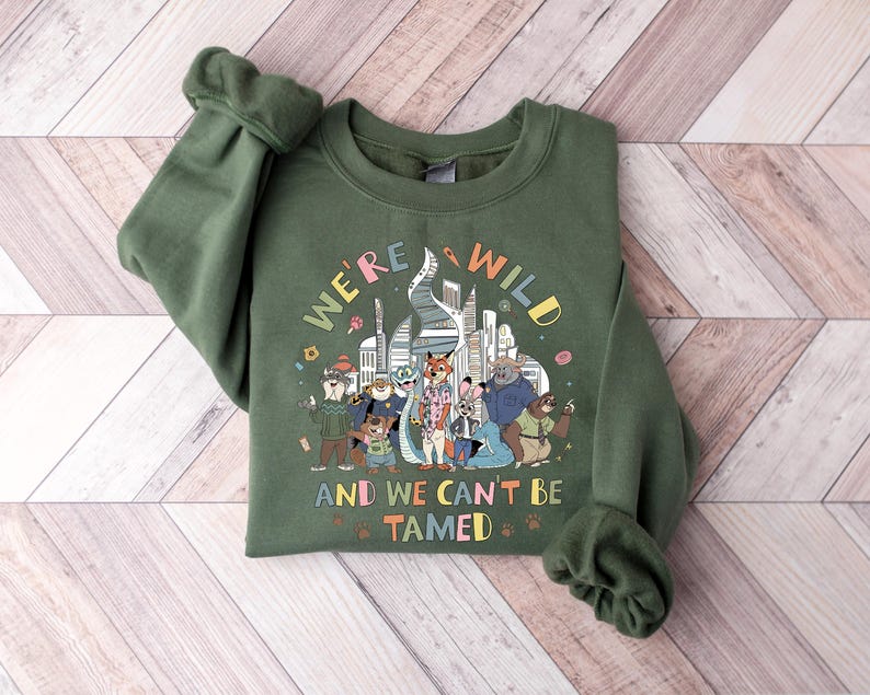 Puede incluir: Una sudadera verde oliva con el texto "WE'RE A WILD AND WE CAN'T BE TAMED" y un gr&aacute;fico colorido de animales antropom&oacute;rficos frente a un horizonte urbano. La sudadera tiene mangas largas con pu&ntilde;os acanalados.