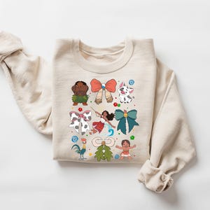 May include: A beige sweatshirt featuring colorful cartoon characters and bows. The design includes a rooster, a pig, and a girl with a bow. The sweatshirt is made of a soft material and has a crew neck.