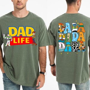 K&ouml;nnte beinhalten: Olivgr&uuml;nes T-Shirt mit dem Text "DAD LIFE" in Gelb und Rot, mit den Zeichentrickfiguren Buzz Lightyear und Woody. Auf der R&uuml;ckseite des Shirts steht "DADA DADA DADA" in einer farbenfrohen, verspielten Schrift.