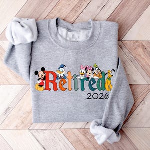 Może przedstawiać: Szara bluza z napisem "Retired" w kolorowych literach, z postaciami Disneya Myszki Miki, Kaczora Donalda, Kaczki Daisy, Myszki Minnie, Goofy'ego i Pluto. Rok "2026" jest wydrukowany poniżej.